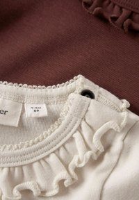 Zwei Kleidungsstücke: ein weißer Stoff mit gerafftem Ausschnitt und ein brauner Stoff darunter. Etiketten zeigen die Größe 4-6M an, mit einem schwarzen Druckknopfverschluss.