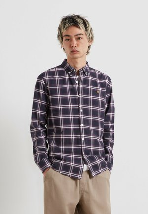 Young man with black and blonde hair wears a dark purple checkered button-up shirt and beige pants, standing against a plain light background.