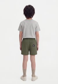 Παιδί με σκούρα μαλλιά στέκεται με την πλάτη γυρισμένη, φοράει γκρι μπλούζα, ελαιχρώμα σορτς με λογότυπο στην πίσω τσέπη, μπεζ κάλτσες και μπεζ παπούτσια.