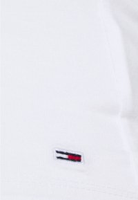 T-shirt bianco in cotone con texture a costine, caratterizzato da un piccolo logo ricamato con strisce rosse, bianche e blu sull'orlo.