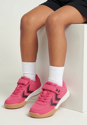 Jambes d'enfant portant des chaussettes blanches et des baskets rose vif avec des bandes chevrons noires, assises sur un bloc blanc sur fond blanc.