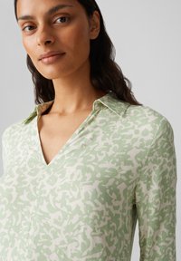 Haut vert clair avec un col en V, présentant un motif blanc abstrait texturé, créant un effet de design superposé.