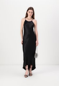 Svart satengslipdress med tynne stropper, høy-lav kant, jevn tekstur, kombinert med svarte hæler med stropper og en liten strukturert håndveske.