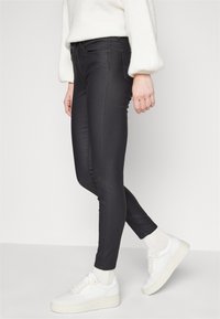 Jeans negros ajustados hechos de material elástico, con un corte slim, bolsillos frontales y detalles de costura sutiles, combinados con zapatillas blancas.