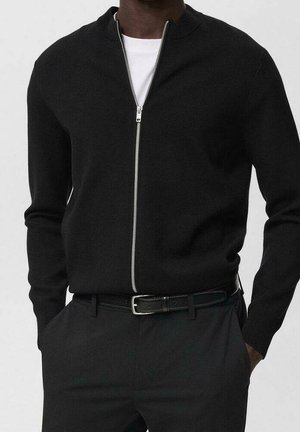 Homme portant un cardigan noir zippé par-dessus une chemise blanche avec un pantalon noir et une ceinture en cuir noire, les mains partiellement dans les poches.