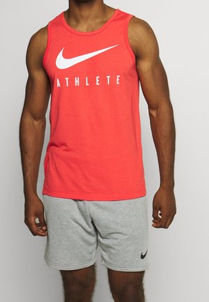 Gespiervd persoon draagt een rood Nike tanktop met de tekst "ATHLETE" en grijze Nike shorts, staand tegen een effen lichte achtergrond.