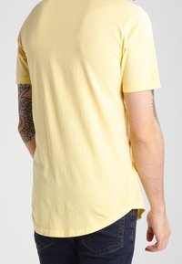 Hombre con una camiseta de manga corta amarilla clara y jeans oscuros, con tatuajes visibles en ambos antebrazos, de pie con la espalda facing.