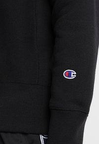 Felpa nera con un tessuto tessuto, polsini a costine e un piccolo logo ricamato in bianco, rosso e blu sulla manica sinistra.