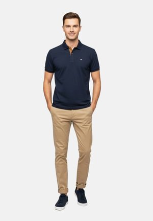 Hombre de pie, vestido con una camiseta polo azul marino, chinos beige remangados y zapatillas azul marino, con las manos en los bolsillos, sonriendo a la cámara.