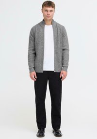 Man staat, draagt een grijze gebreide cardigan met ritssluiting over een wit T-shirt, zwarte broek en zwarte nette schoenen, tegen een effen witte achtergrond.