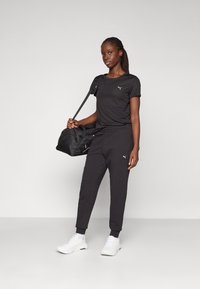Schwarzes Sportoutfit mit einem eng anliegenden T-Shirt und schmal zulaufenden Jogginghosen mit Logo. Das Model trägt eine schwarze Sporttasche und weiße Turnschuhe.