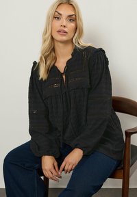 Mustakalla toppi, jossa on teksturoitu kangas, rypytettyjä yksityiskohtia, avainreikäpääntie nauhoilla, sekä brodeeratut yksityiskohdat hihoissa ja helmassa.