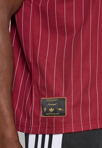 Mørkerød stribet Arsenal-trøje med sort og guld label nær kanten, båret med sort og hvid Adidas stribede bukser.