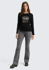 Woman standing wearing black long-sleeve "beautiful" print top, gray flared jeans, and black platform shoes on plain light background.