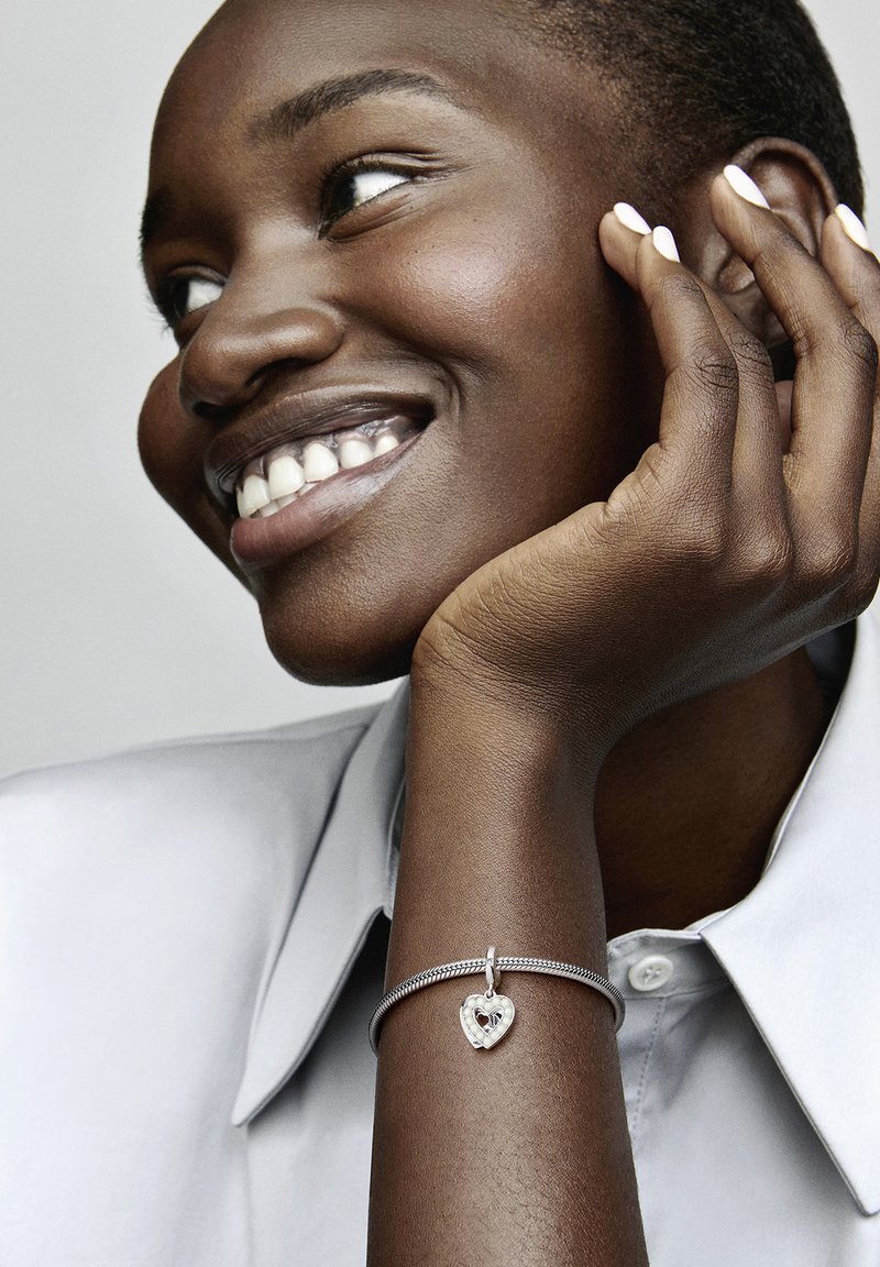 Bracciale d'argento con un ciondolo a forma di cuore e una piccola pietra preziosa incastonata. Il modello indossa una camicia grigia chiaro con bottoni.