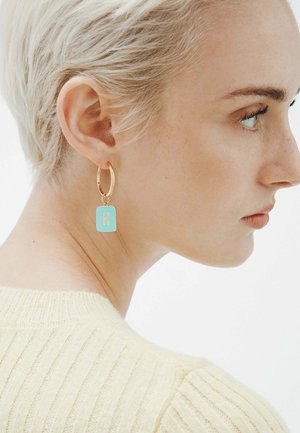Pendientes de aro dorados con un charm rectangular azul claro que tiene la letra "H". La persona que los lleva tiene el pelo rubio corto y viste un suéter de punto amarillo claro.