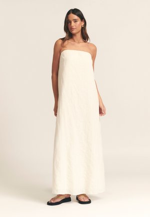 Robe sans manches, longue jusqu'au sol, en tissu crème clair avec une texture verticale. Associée à des sandales plates, le design est simple et élégant.