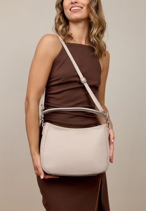 Lächelnde Frau in braunem ärmellosen Kleid, die eine beigefarbene Ledertasche mit verstellbarem Schultergurt vor neutralem Hintergrund hält.