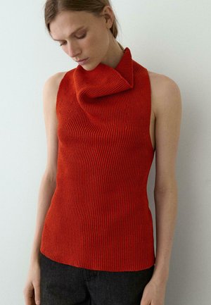 Femme portant un haut rouge sans manches en maille côtelée avec col bénitier asymétrique, debout contre un mur clair, regardant vers le bas.