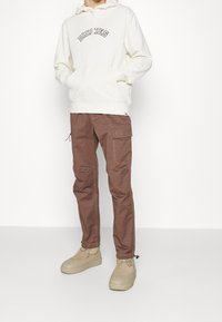 Sudadera con capucha crema con el texto "DICKIES", combinada con pantalones cargo marrones que cuentan con múltiples bolsillos y zapatos de plataforma beige.
