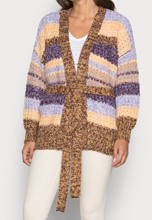 Femme portant un cardigan en tricot épais à rayures violettes, jaunes et marron, ceinturé à la taille, associé à un pantalon blanc et une chemise blanche.
