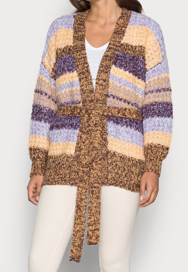 Femme portant un cardigan en tricot épais à rayures violettes, jaunes et marron, ceinturé à la taille, associé à un pantalon blanc et une chemise blanche.