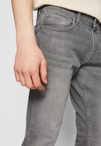 Sive jeans iz džinsa z ozkim krojem, s subtilnim zbledelostjo, petimi žepi in vidnimi šivi. Stranski pogled, ki prikazuje področje bokov.