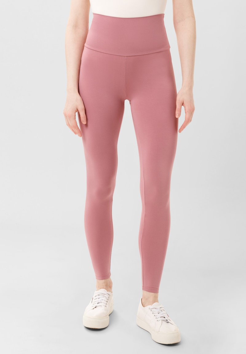Roze high-waisted leggings van elastische stof, met een gladde textuur en een strakke pasvorm, gecombineerd met witte veterschoenen.