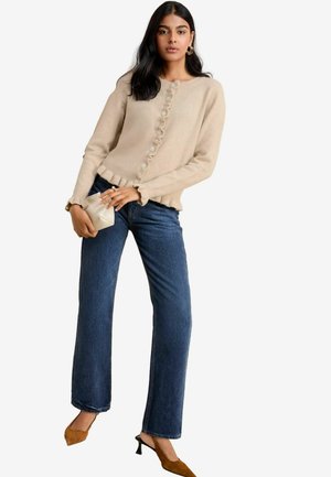 Beige Strickjacke mit Knopfverschluss, Rüsche an den Bündchen, kombiniert mit dunkelblauen, geraden Jeans und braunen, höheren Mules.