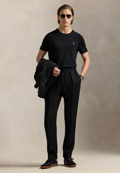 Mann trägt schwarze Sonnenbrille, schwarzes T-Shirt mit Logo, plissierte schwarze Hose, hält eine schwarze Jacke, steht vor schlichtem Hintergrund.