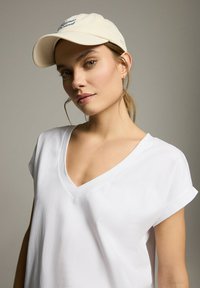 Biała bawełniana koszulka typu v-neck z podwiniętymi krótkimi rękawami; kremowa czapka z niebieskim haftowanym logo; ciepły odcień skóry i neutralne tło.