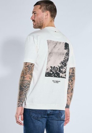 Mann mit Tattoos auf beiden Armen, der ein weißes T-Shirt mit schwarz-weißem Naturmotiv und Text auf dem Rücken trägt, kombiniert mit blauen Jeans.