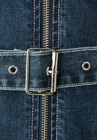 Szczegół kurtki dżinsowej z widocznym złotym zamkiem błyskawicznym, srebrną prostokątną klamrą, niebieskim materiałem z białymi przeszyciami oraz oczkami do zapinania.