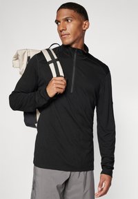 Top preto de manga longa com fecho, apresentando uma textura suave e um design ajustado. Usado com uma mochila que exibe alças claras.