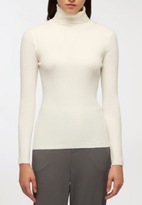 Dopasowany kremowy golf z długim rękawem, o prążkowanej teksturze i gładkim wykończeniu. Noszony z szarymi spodniami, podkreślającymi jego sylwetkę.