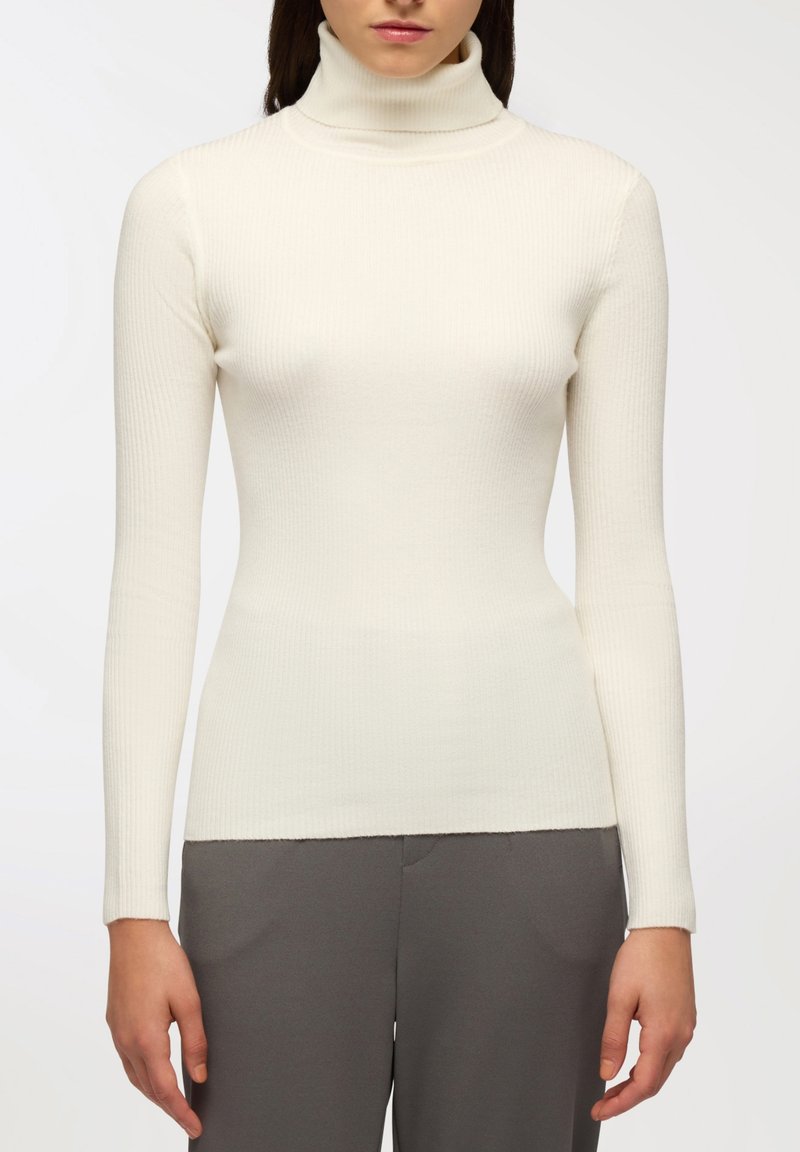 Dopasowany kremowy golf z długim rękawem, o prążkowanej teksturze i gładkim wykończeniu. Noszony z szarymi spodniami, podkreślającymi jego sylwetkę.