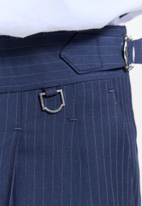 Pantalon bleu marine à fines rayures avec un détail de boucle métallique à la ceinture, porté avec un haut blanc.