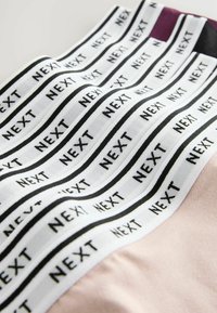 Krupni plan bijelih elastičnih trakica s crnim natpisom "NEXT" ponavljanim, pričvršćenim za tkaninu u ružičastoj, crnoj i ljubičastoj boji.