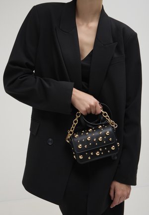 Sac à main noir avec des clous dorés, en forme rectangulaire, avec de courtes poignées et une bandoulière en chaîne. Porté avec un blazer noir surdimensionné.