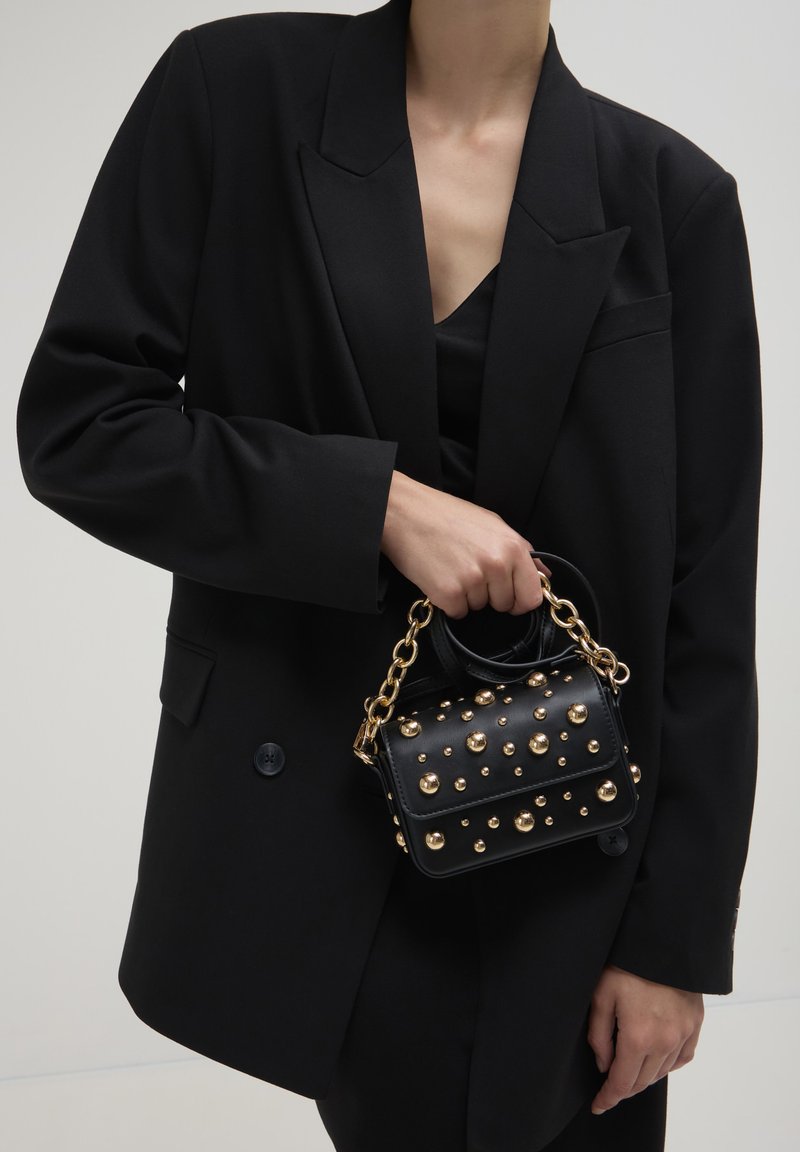 Borsa nera con borchie dorate, forma rettangolare, manici corti e una tracolla a catena. Indossata con un blazer oversize nero.