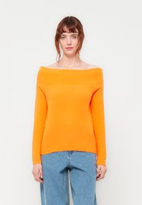 Pulover v oranžni barvi z odkrito ramena, izdelan iz pletene tkanine, s dolgimi rokavi in prilegnjenim krojem, kombiniran z svetloblue jeans hlačami.