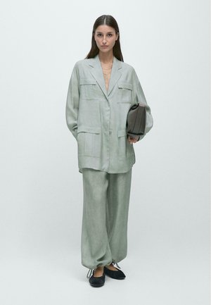 Frau steht in einem hellgrauen, übergroßen Anzug mit großen Taschen, hält eine dunkelgraue Clutch und trägt schwarze Ballerinas vor einfarbigem Hintergrund.