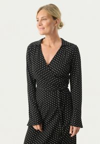 Schwarzes Wickelkleid mit langen Ärmeln, das weiße Pünktchen, einen Kragen und eine gebundene Taille aufweist, aus leichtem Stoff gefertigt.