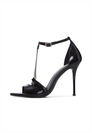 Czarne lakierowane sandały na wysokim obcasie z odkrytym noskiem, paskiem wokół kostki z ozdobą z srebrnego łańcuszka oraz obcasem typu szpilka.