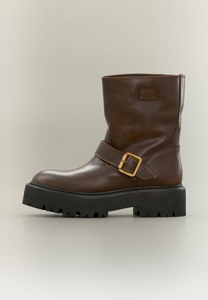Brown leather ankle boot with thick black lug sole and gold buckle strap on the side against neutral background.
