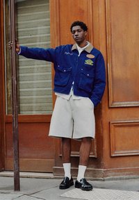 Mladý muž v modré bundě, béžových krátkých kalhotách, bílých ponožkách a černých botách stojí venku a drží tyč u dřevěných dveří.