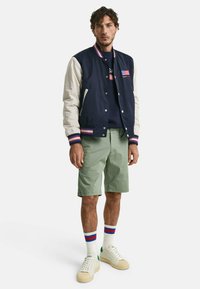 Marine- og kremfarget bomberjakke med stripete mansjetter, grønne shorts, hvite sneakers med grønne detaljer, og stripete sokker.