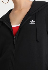 Svart hoodie i mjukt material med dragkedjestängning, snörning vid huvan och vit Adidas-logotyp på bröstet, med en röd topp under.