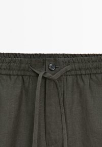 Pantaloni corti verde scuro con coulisse e cintura elastica, dotati di un bottone nero e tessuto testurizzato. Vista ravvicinata dei dettagli della vita.