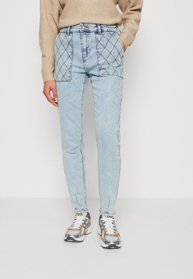 Jean bleu clair avec poche patch matelassée porté avec un pull beige et des baskets blanches, argentées et orange sur fond blanc.